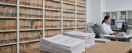Stacks of printed order packets on a desk in front of file shelves, a staff member working in the background